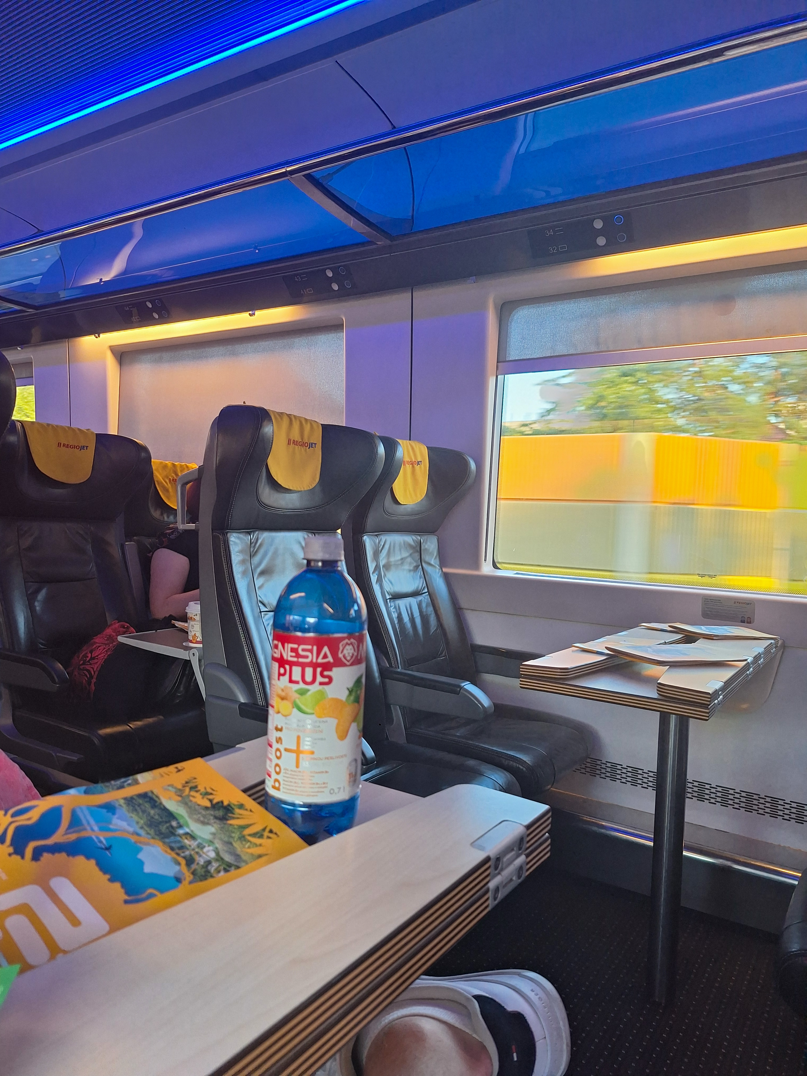 Interior of a RegioJet first class carriage with yellow leather seats, blue mood lighting overhead, a bottle of Vinea Plus on the table and a magazine on the next seat